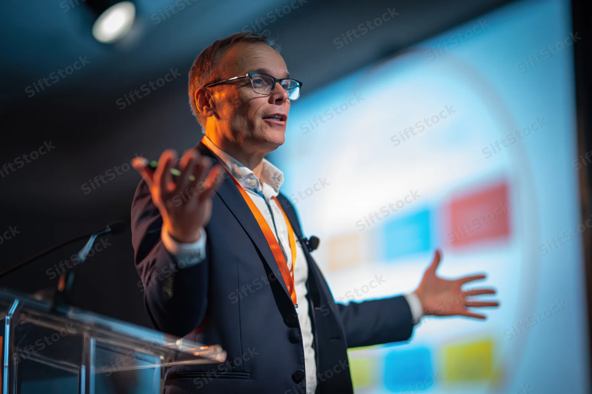 Man in suit gesturing while speaking at podium with projection screen behind him