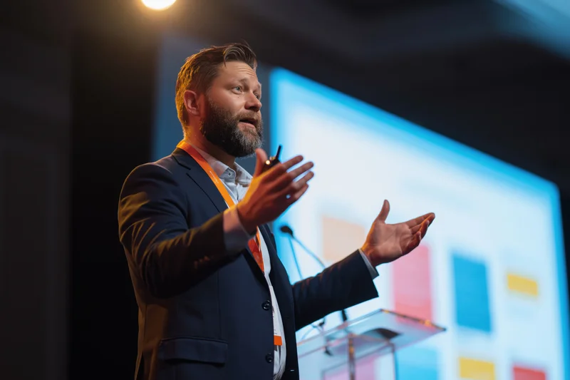 Man in suit gesturing while speaking at a conference stage with a presentation screen behind him
