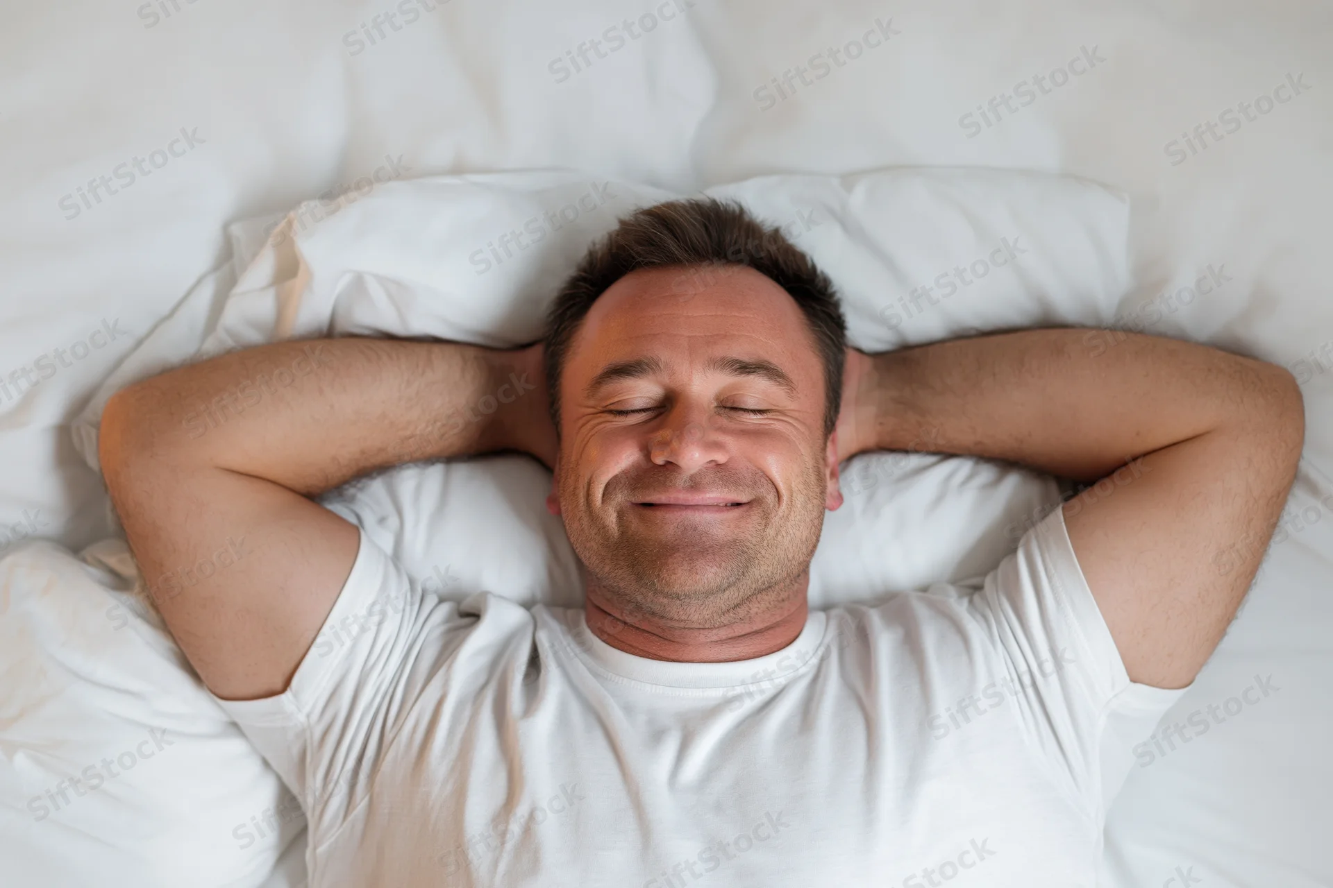 Man lying in bed with hands behind his head smiling, eyes closed, white bedding, overhead view