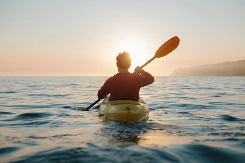 Man paddling a yellow kayak on calm coastal water at sunset