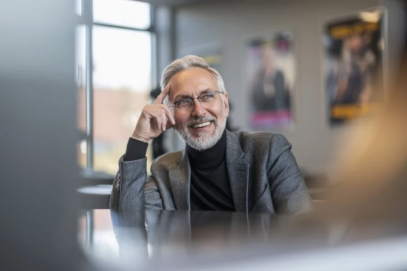 Mature businessman smiling while thinking at office desk with hand on head