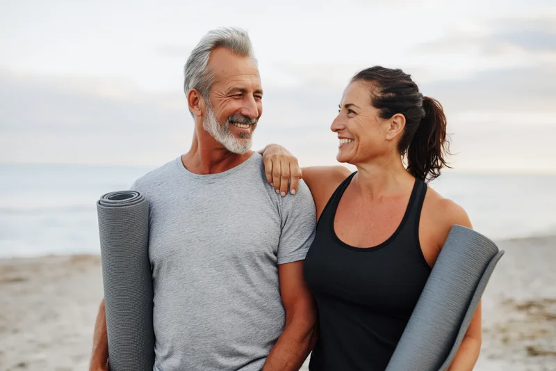 Mature couple smiling at each other on beach holding yoga mats