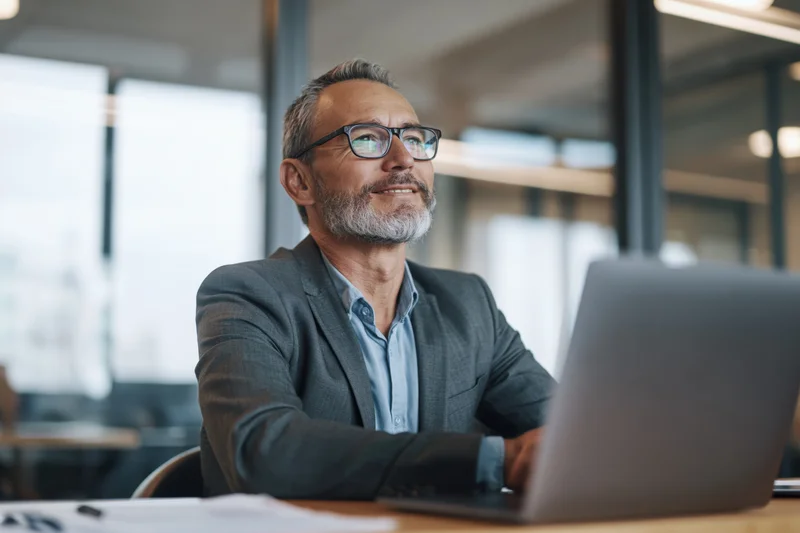 Middle-aged businessman with grey-flecked beard and glasses smiling while working on laptop at office desk