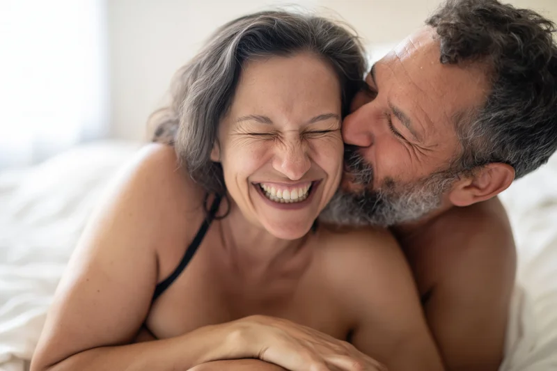 Middle-aged couple embracing on bed, man kissing woman’s cheek as she laughs joyfully