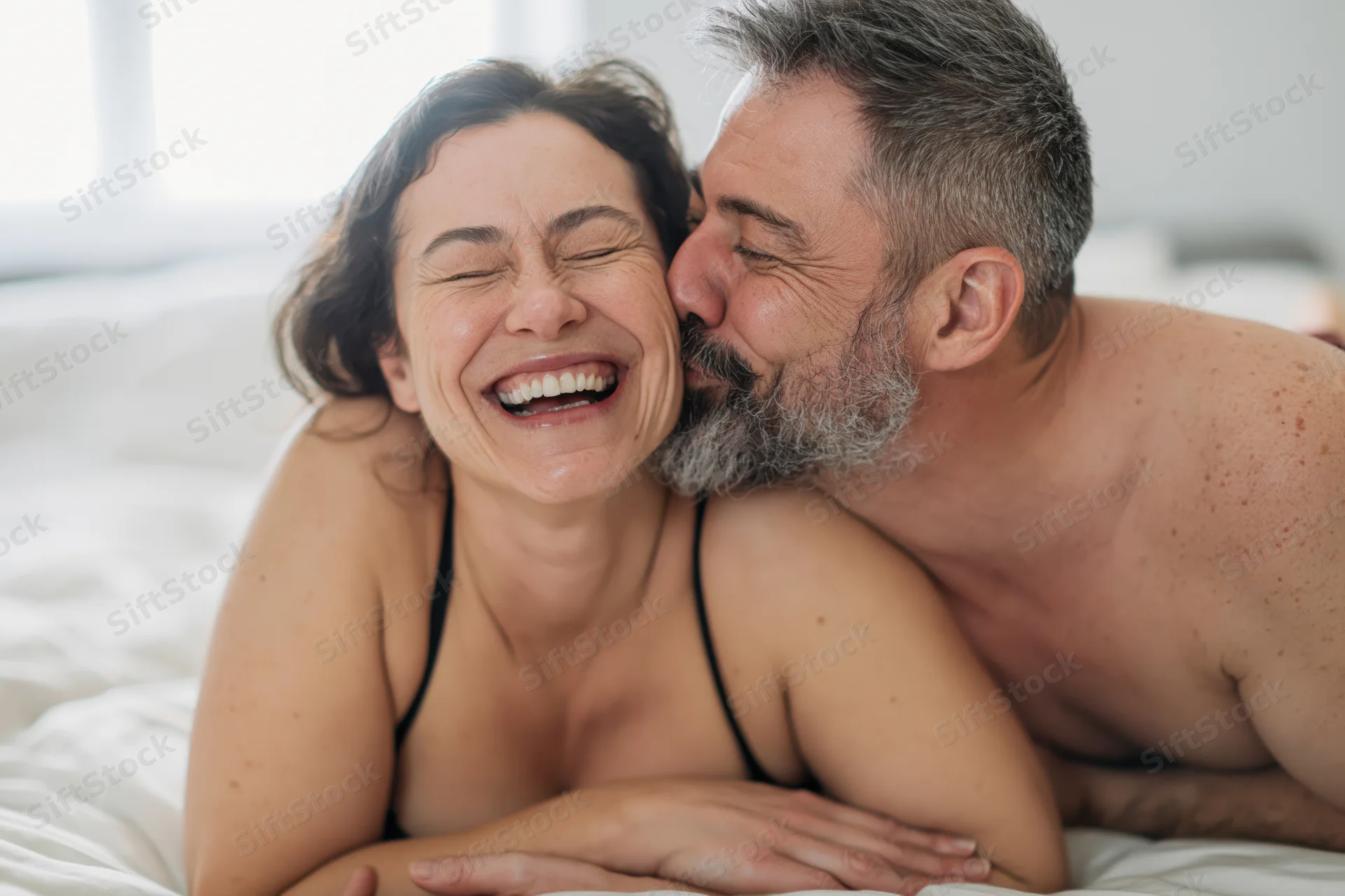 Middle-aged couple laughing and kissing in bed, woman laughing with eyes closed, man embracing her from behind
