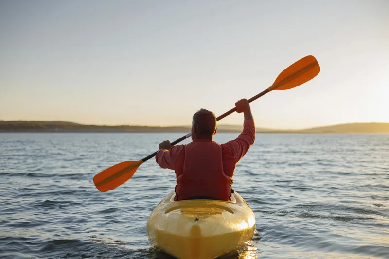 Man paddling a yellow kayak on calm water at sunset