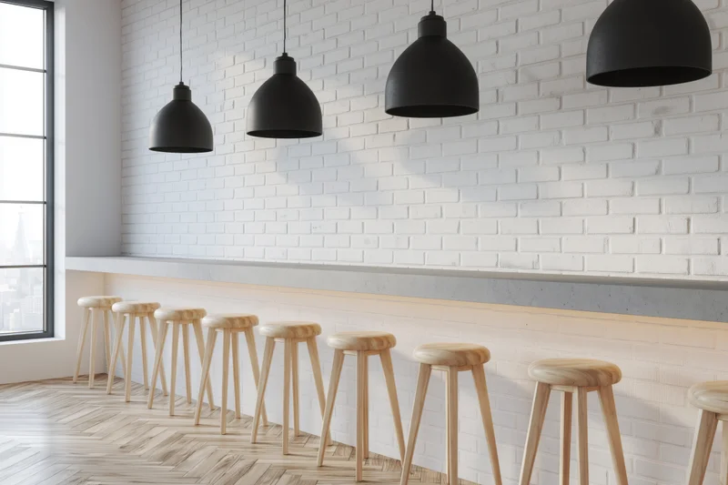 Modern minimalist bar interior with white brick walls, wooden stools, black pendant lights, and concrete counter