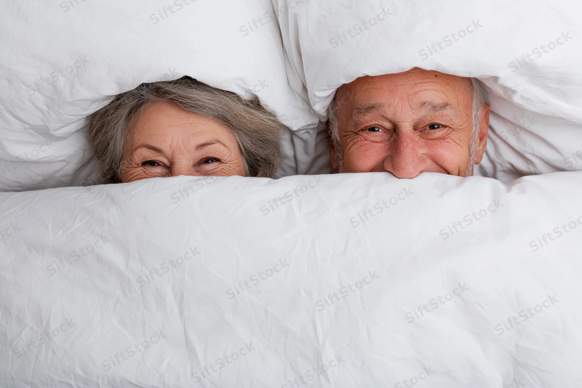 Senior couple peeking playfully over white blanket in bed