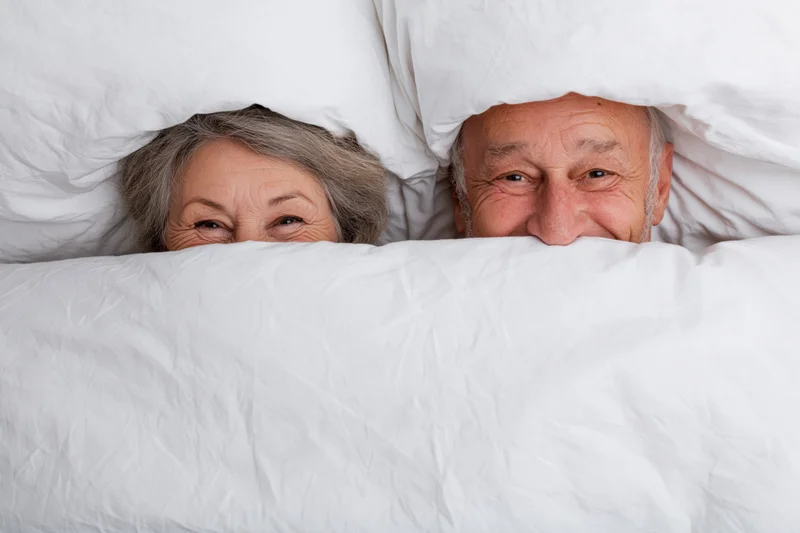 Senior couple peeking playfully over white blanket in bed