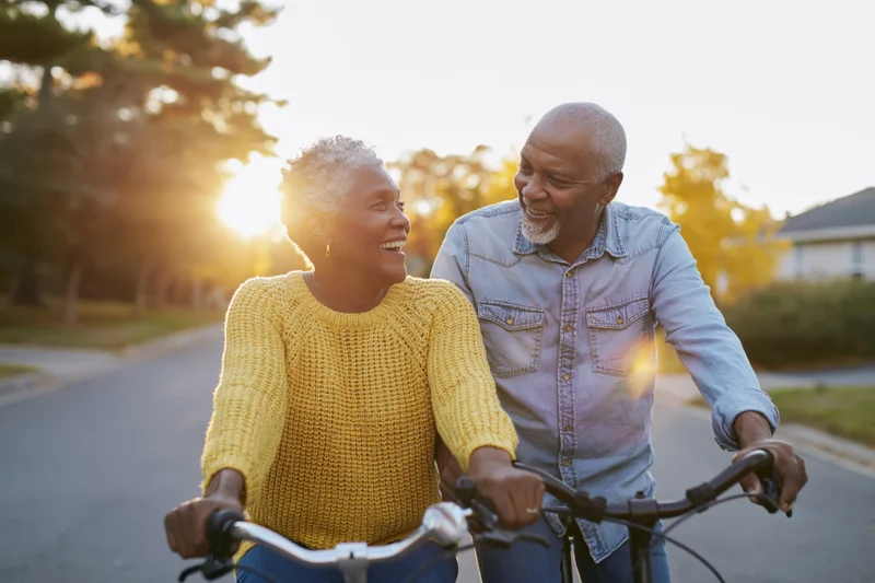 Senior black couple smiling while riding bicycles down a quiet suburban street at golden hour