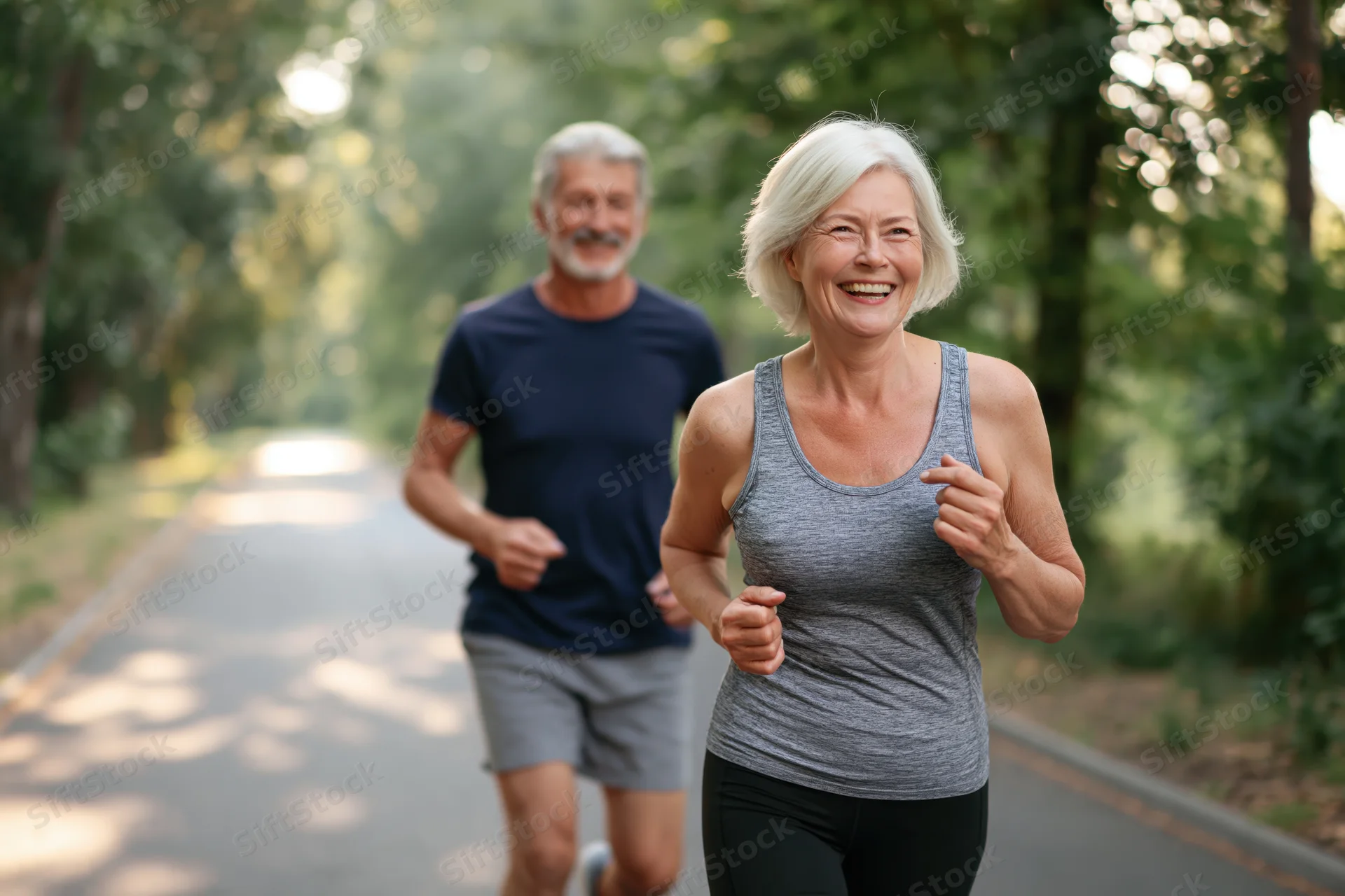 Senior woman jogging with man on tree-lined park path, both smiling and active in athletic wear