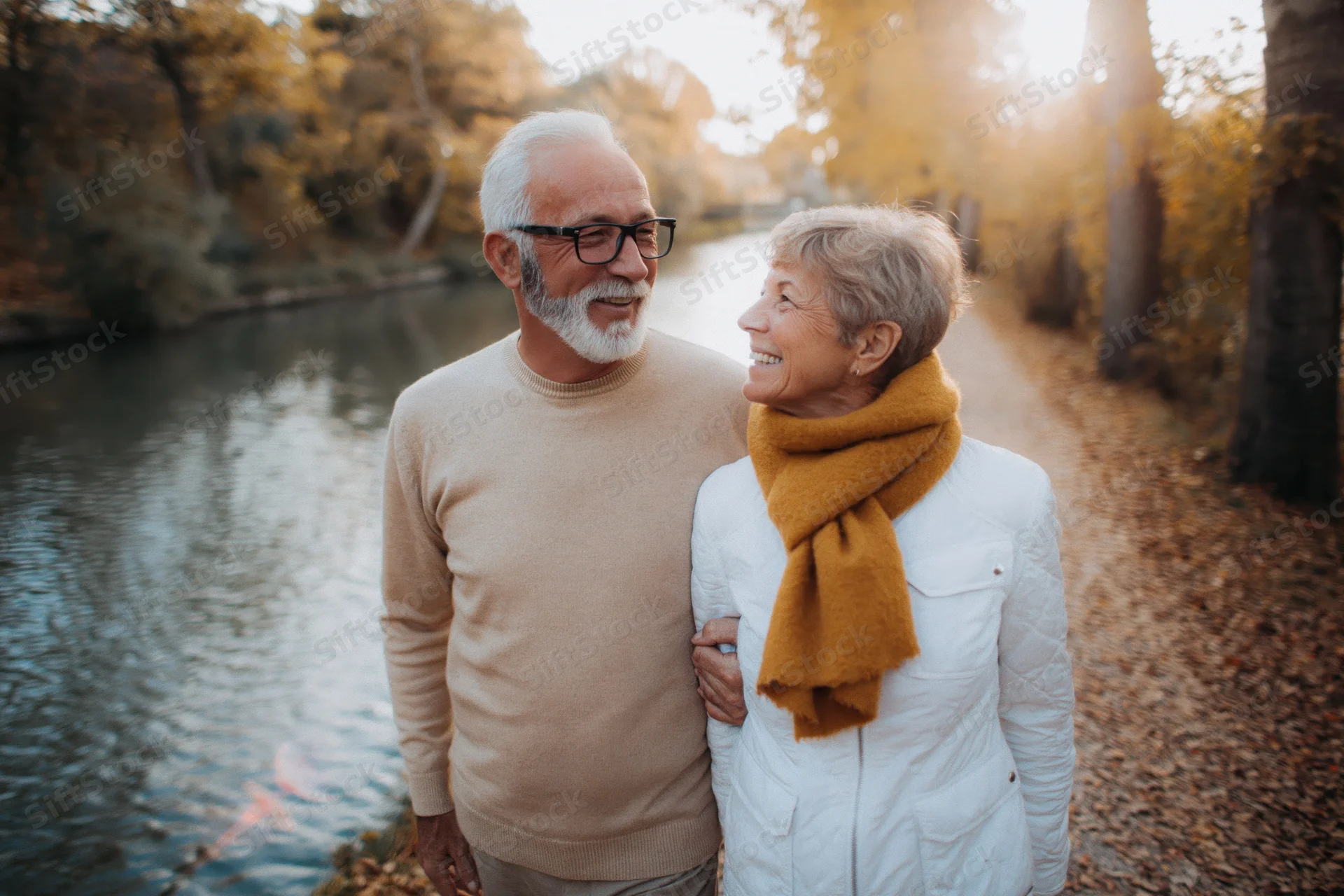 Senior couple walking arm in arm along a leaf-covered path beside a river in autumn park