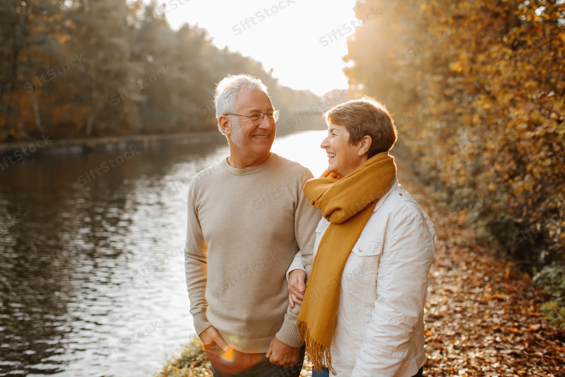 Senior couple smiling and holding hands while walking along a leaf-covered riverbank in autumn
