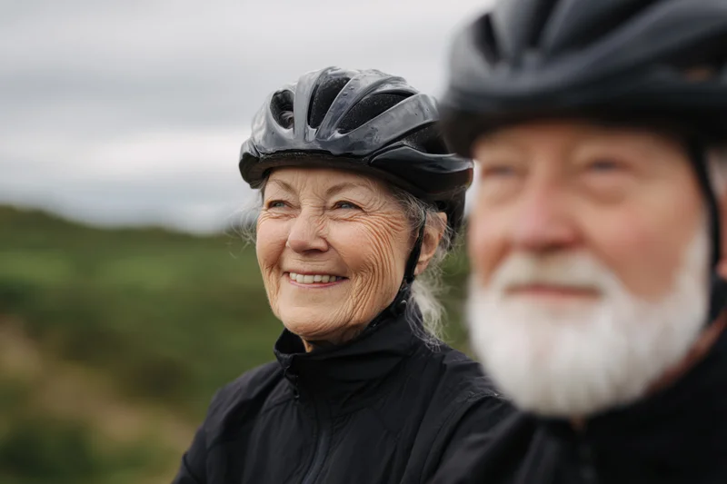 Senior woman and man wearing bicycle helmets smiling outdoors in a green landscape