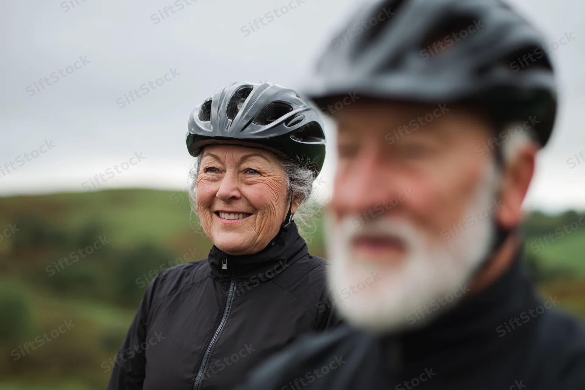 Senior woman smiling while standing outdoors with a man in cycling gear, both wearing helmets in a green countryside setting