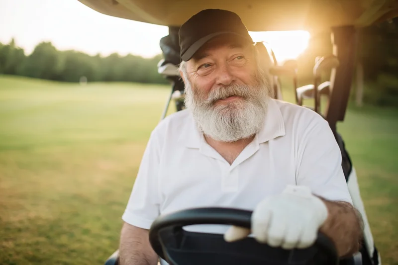 Senior man with grey beard driving golf cart on course at sunset
