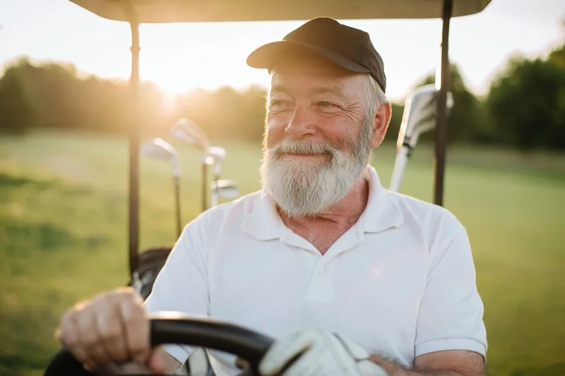 Senior man smiling while driving a golf cart on a green course at sunset