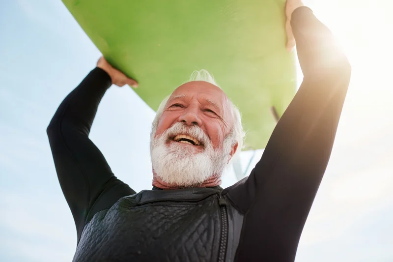 Senior man in wetsuit holding green surfboard overhead, smiling, outdoors