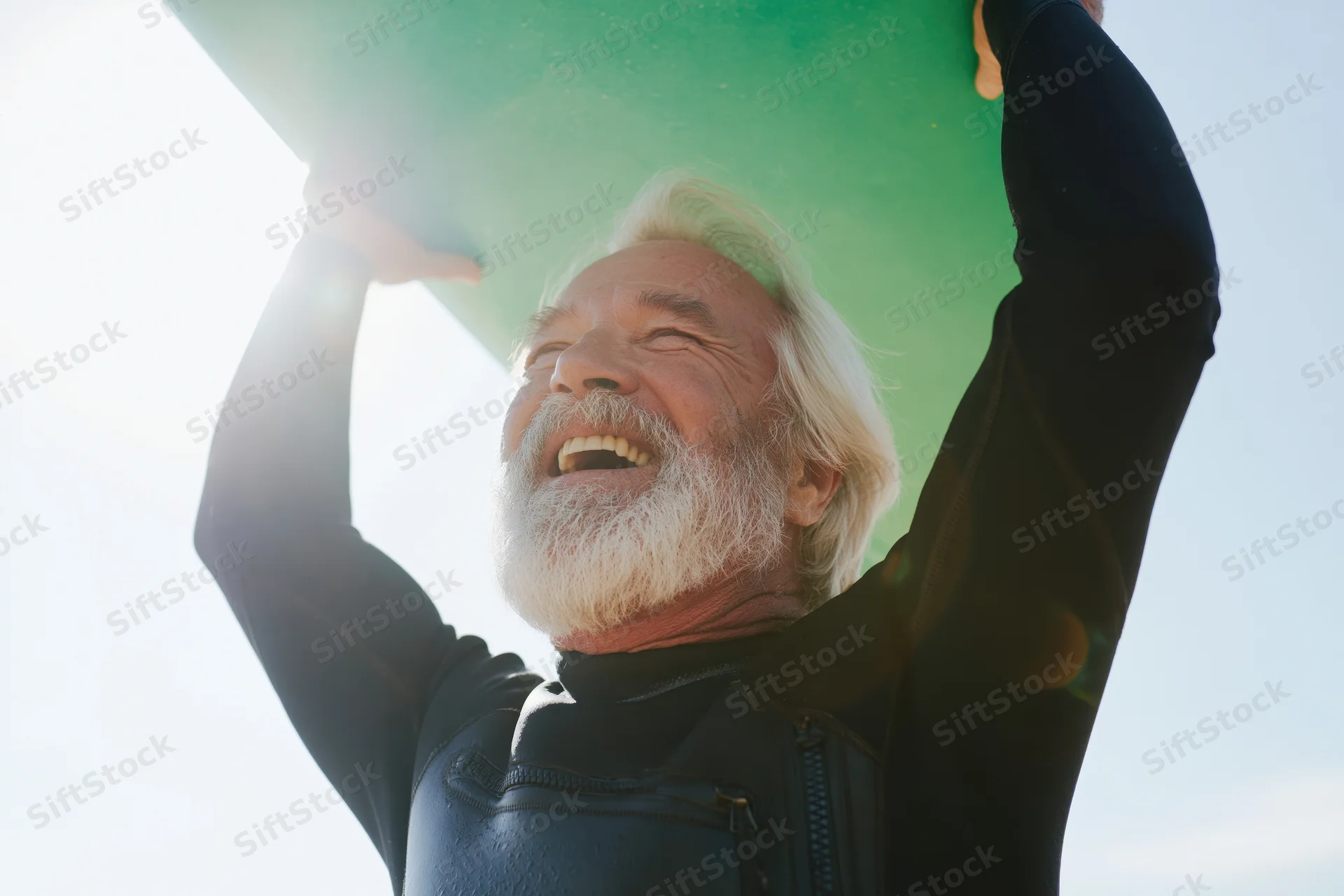 Senior man in wetsuit holding green surfboard overhead, smiling against bright sky