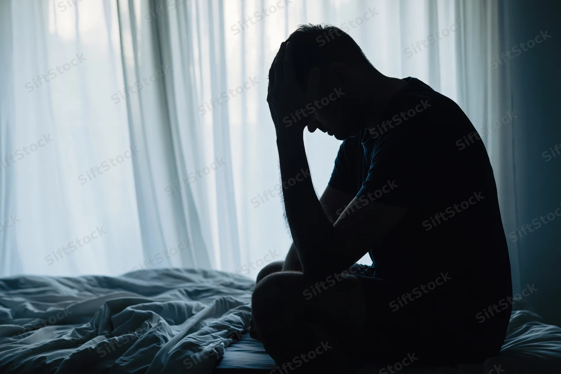 Silhouette of a man sitting on bed edge with head bowed in dimly lit bedroom