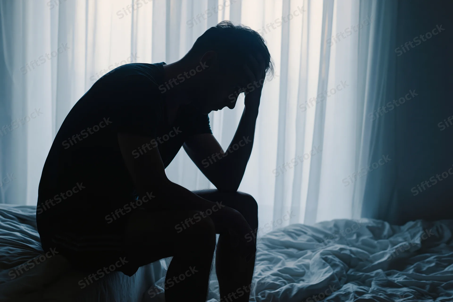 Silhouette of a man sitting on the edge of a bed with head bowed and hand on head in a dimly lit bedroom