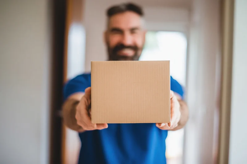 Smiling delivery man holding a cardboard box toward the camera at a doorway