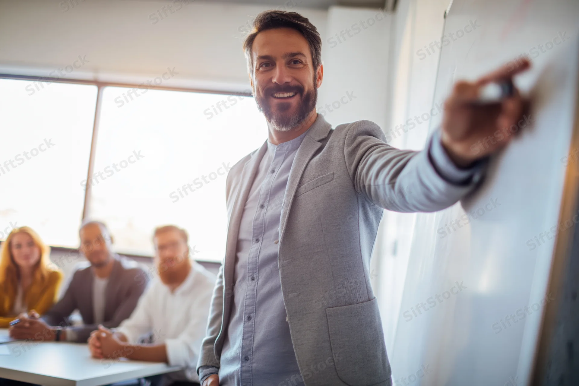 Man presenting at whiteboard in office meeting with team seated at table behind him