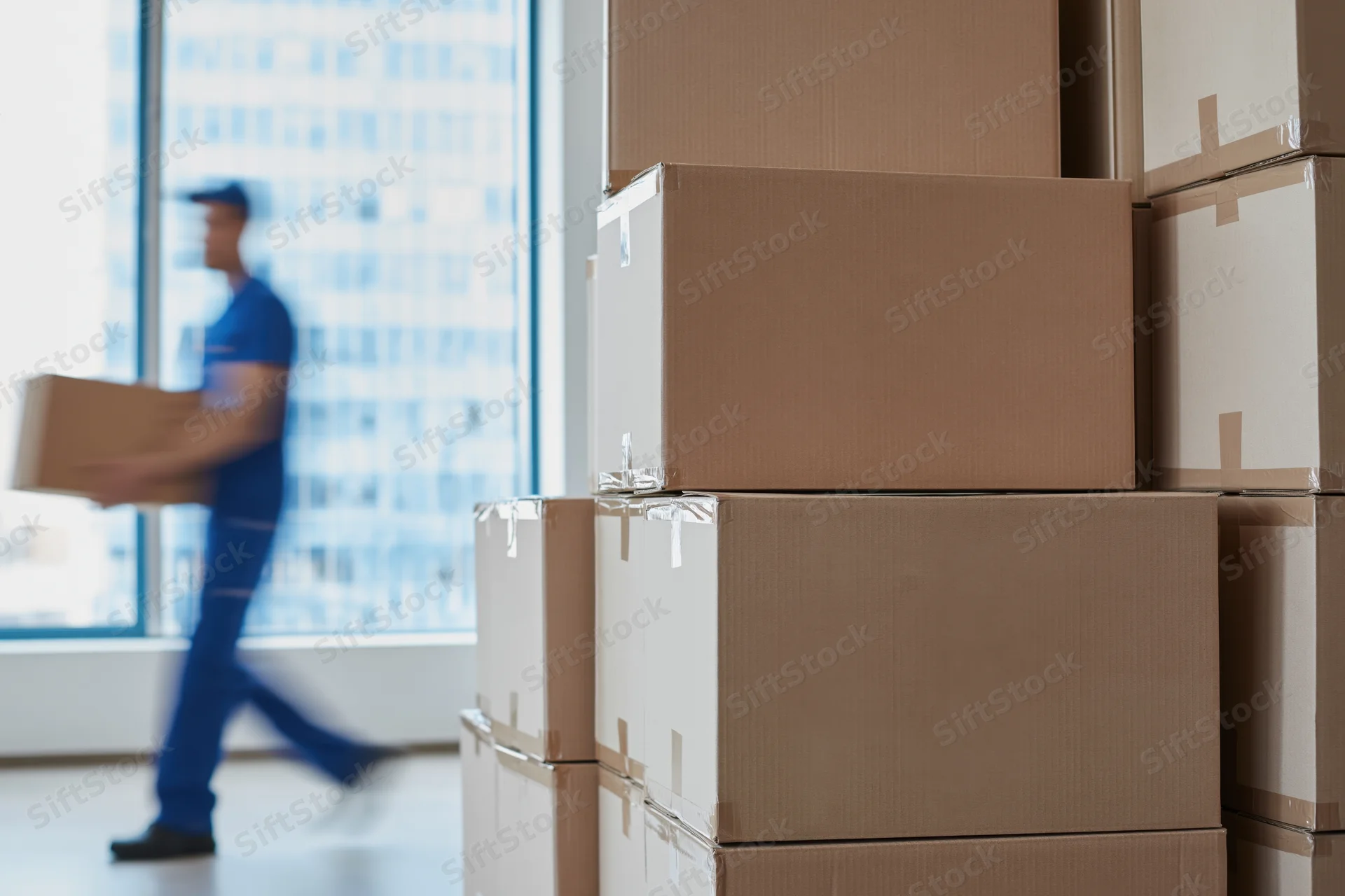 Stacks of cardboard boxes in an office with a delivery worker carrying a box in motion near a window