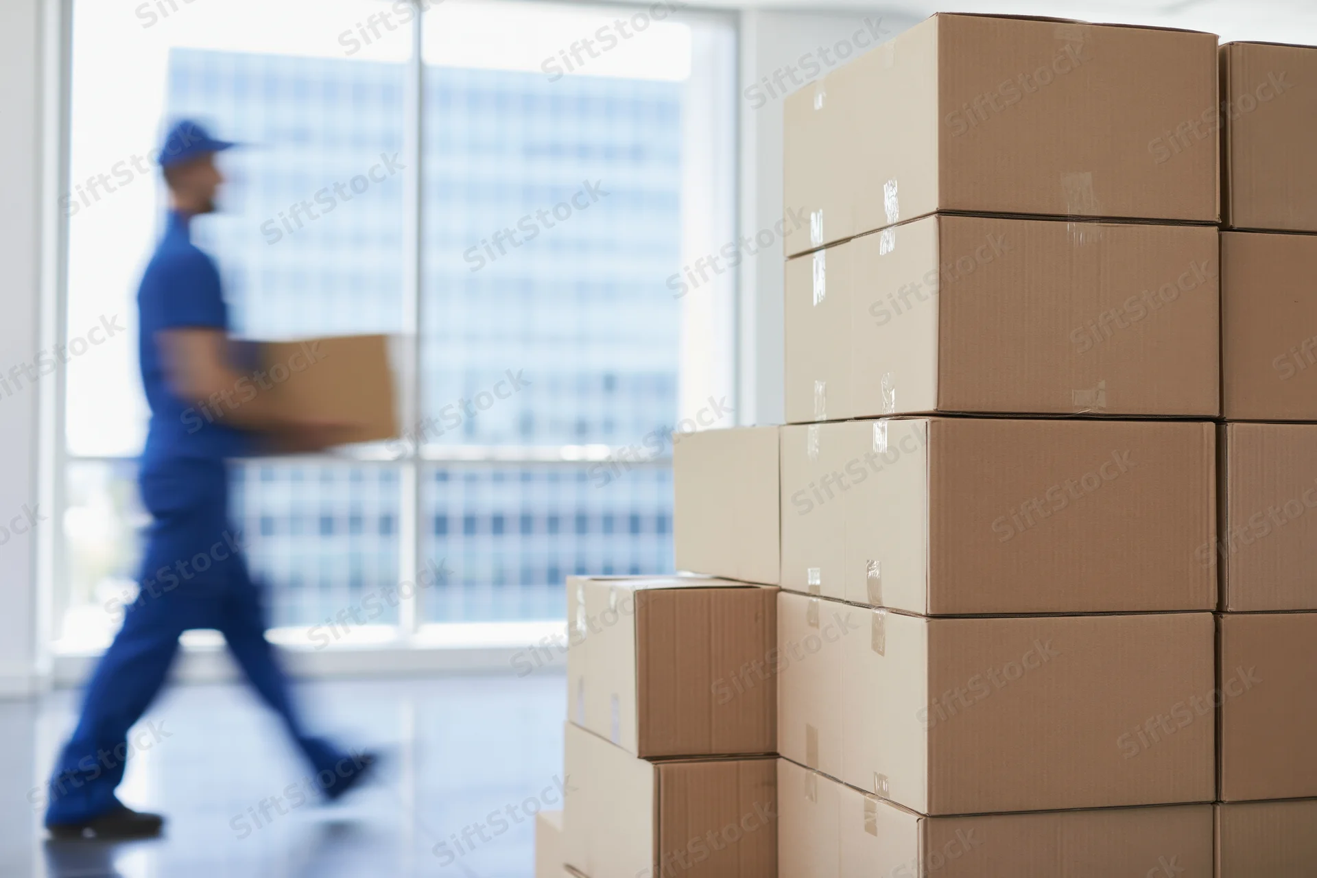 Stacks of cardboard boxes in an office interior with a delivery worker in motion carrying a box near a window