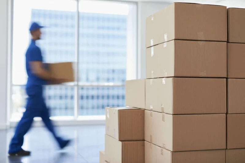 Stacks of cardboard boxes in an office interior with a delivery worker in motion carrying a box near a window