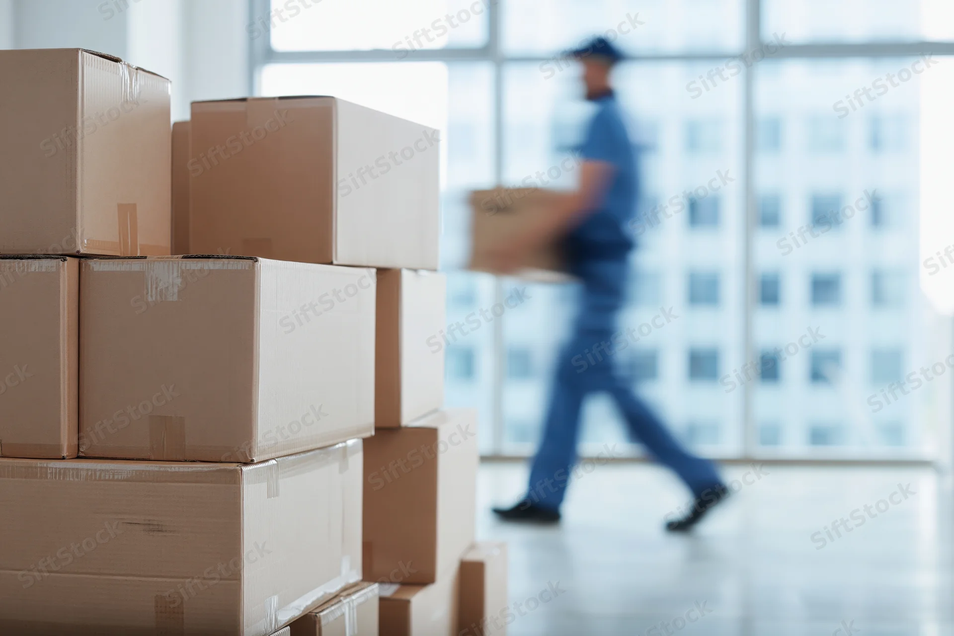 Stacks of cardboard boxes in foreground with delivery worker carrying box in motion behind large windows