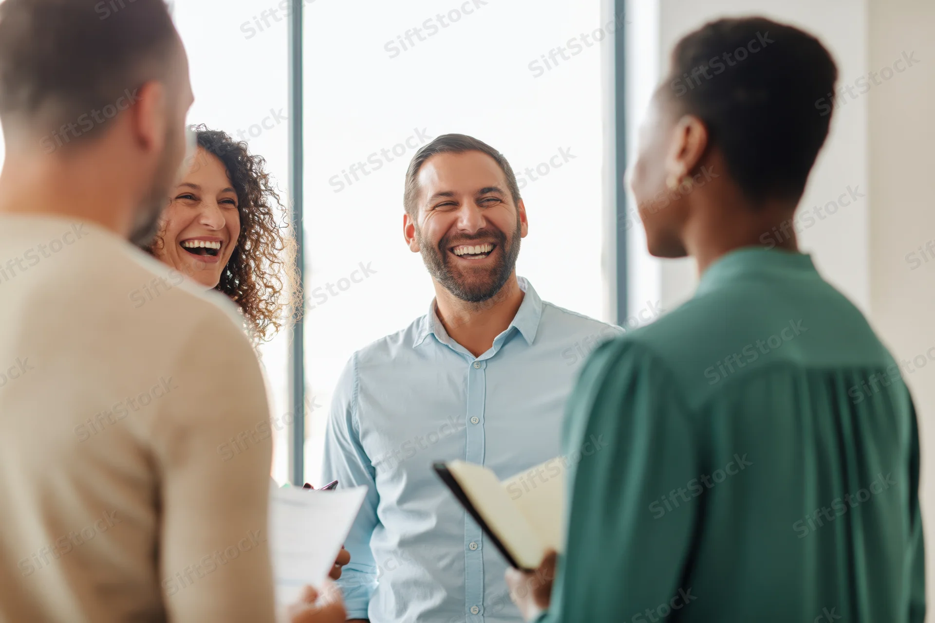 Colleagues laughing during a casual office meeting with papers and notebook in hand