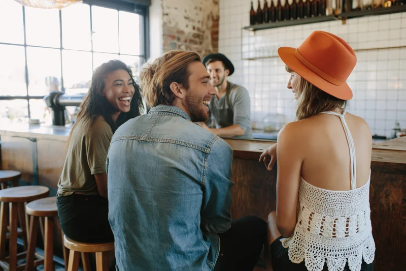 Three friends laughing and talking at a cafe counter while seated on wooden stools