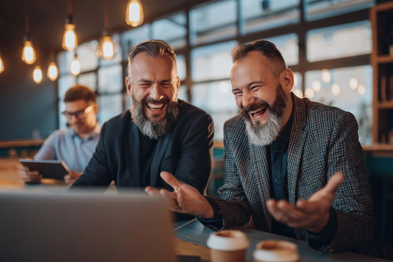 Two bearded men in blazers laughing at a laptop screen in a modern office setting
