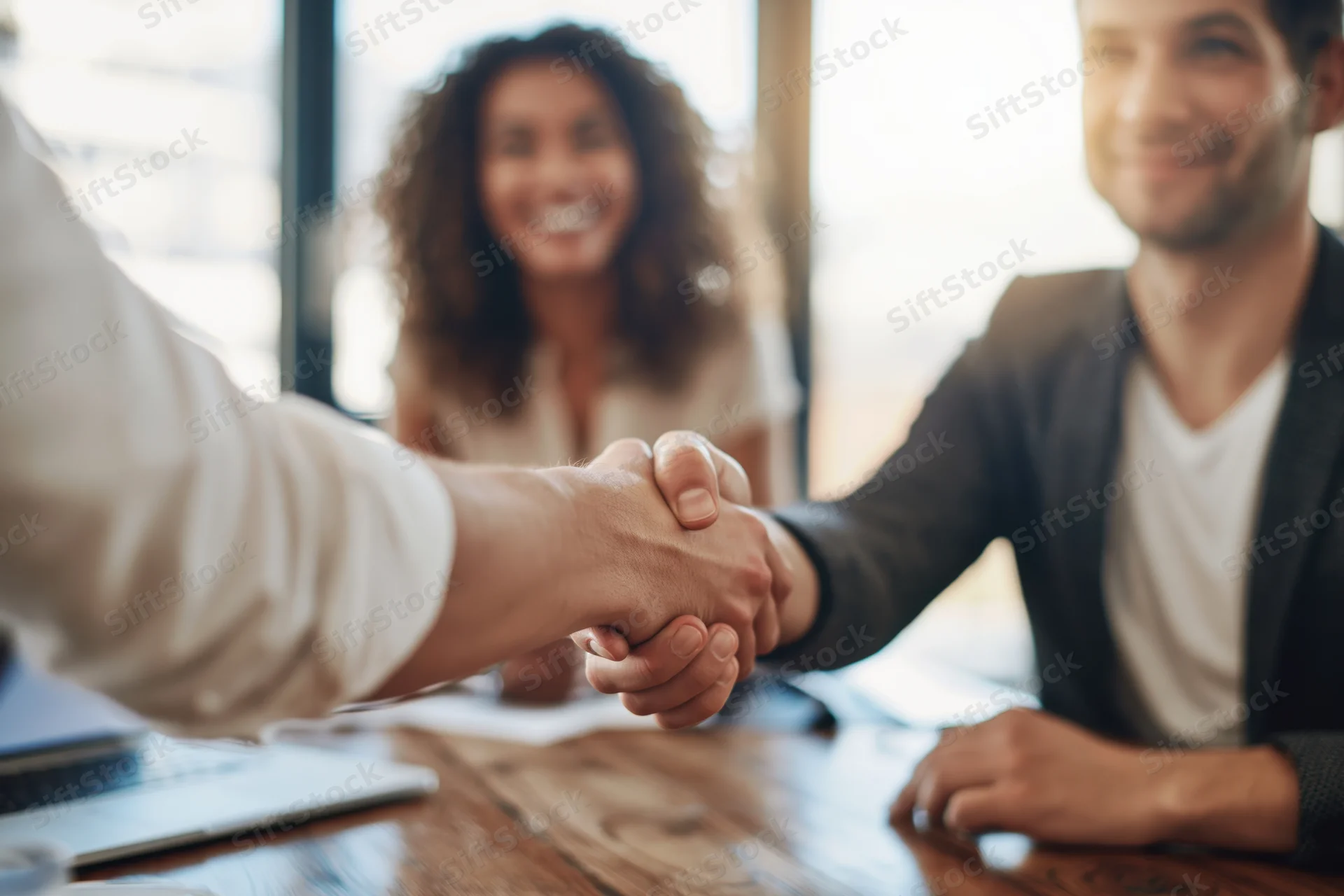 Two business professionals shaking hands across a wooden desk in an office meeting