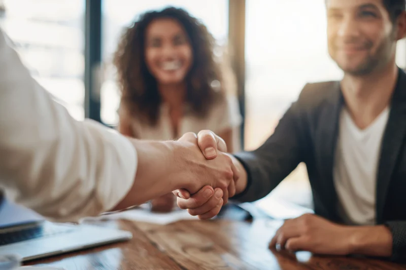 Two business professionals shaking hands across a wooden desk in an office meeting