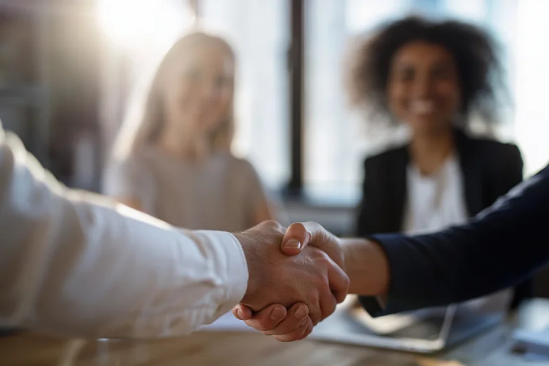 Two business professionals shaking hands across a desk in an office meeting
