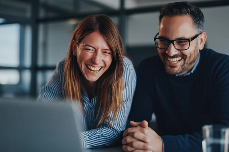 Woman and man laughing together while looking at laptop screen in office setting