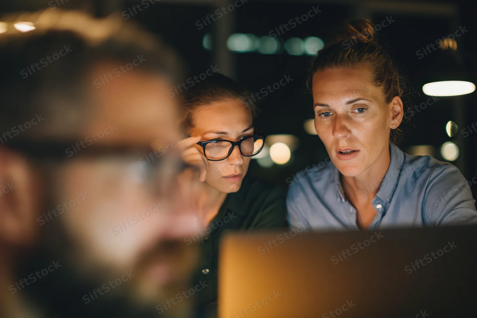 Two coworkers focused on a laptop screen in an office at night