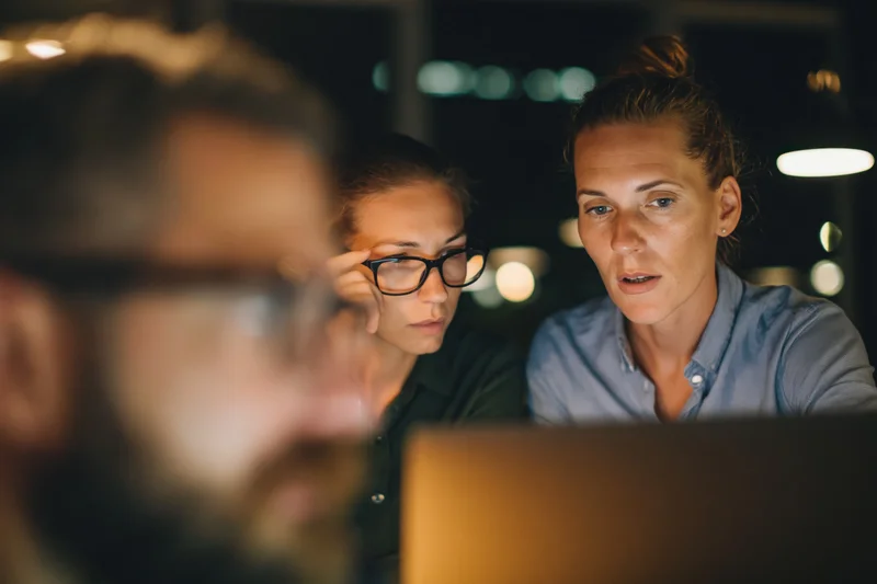 Two coworkers focused on a laptop screen in an office at night
