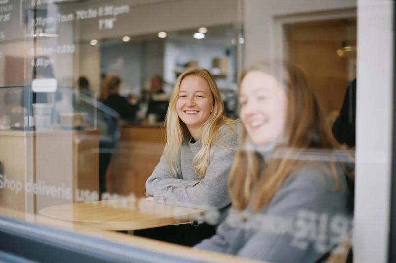 Two women smiling and laughing while sitting at a wooden cafe table viewed through a glass window