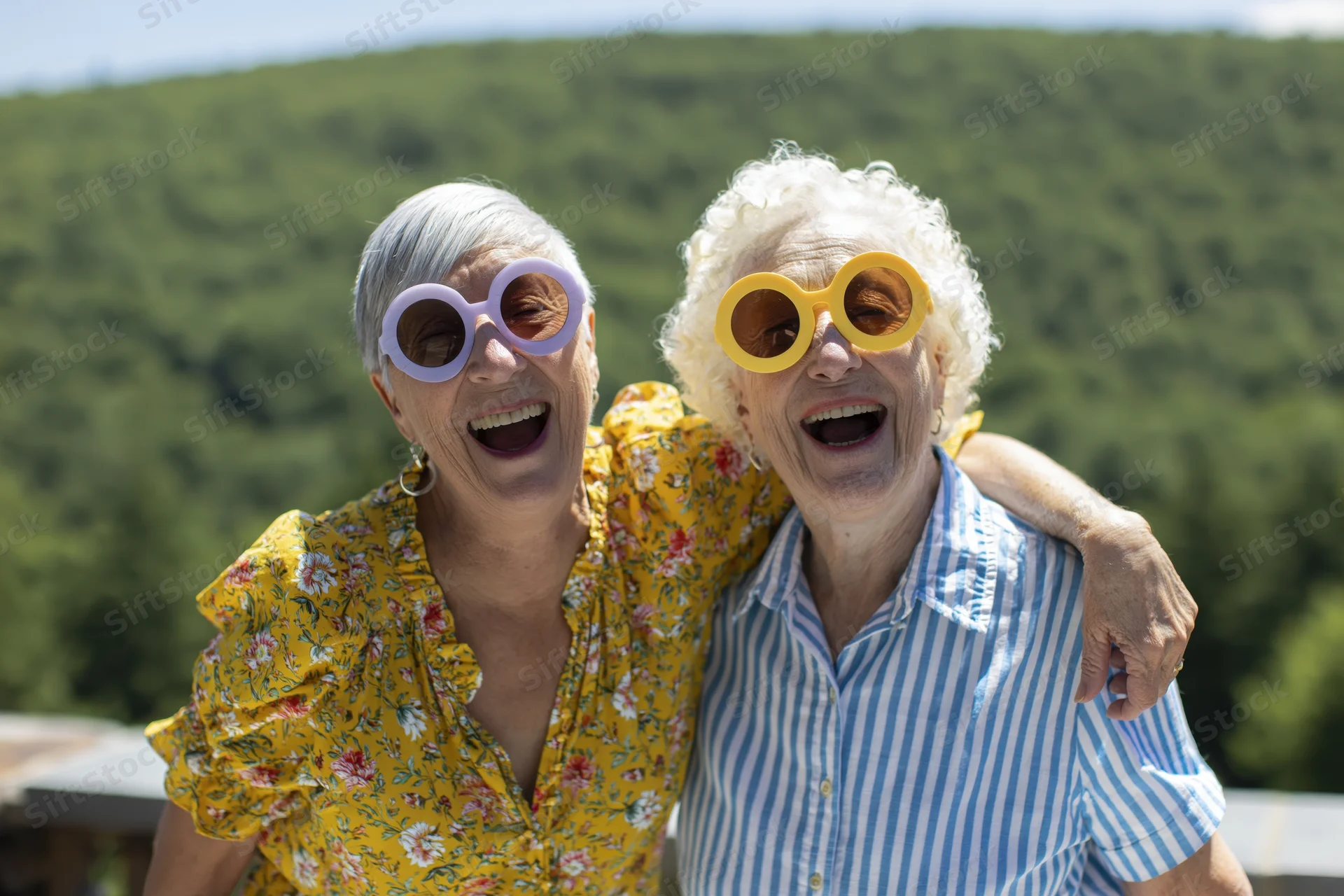 Two senior women laughing together on an outdoor terrace wearing colorful sunglasses
