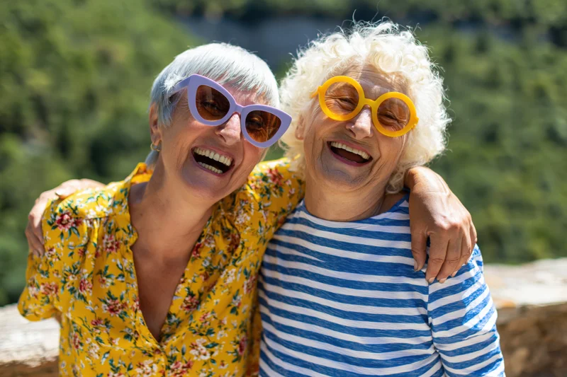 Two senior women laughing together outdoors on a terrace with arms around each other