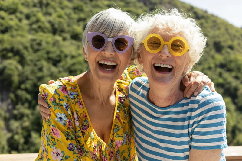 Two senior women laughing together outdoors on a terrace with arms around each other
