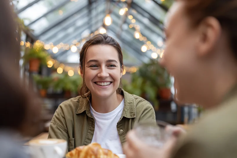 Woman smiling while chatting with friends over coffee at a cafe table in a greenhouse setting