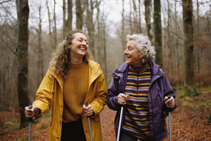 Two women hiking together through an autumn forest laughing while walking on a leaf-covered trail