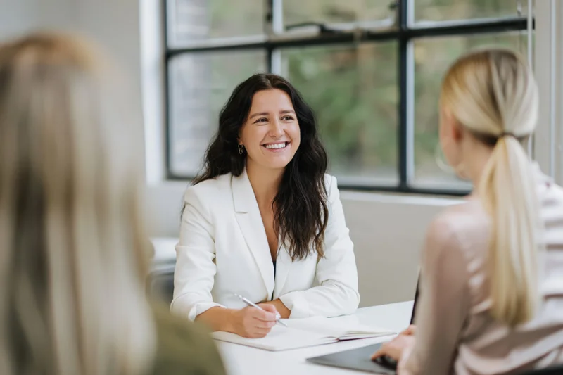 Woman in white blazer smiling during business meeting at desk with colleagues