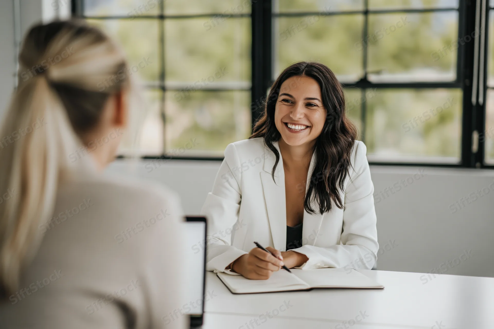 Woman in white blazer smiling during business meeting at white desk with another woman across from her