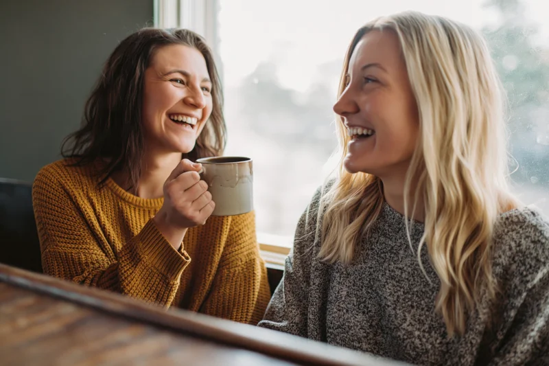 Two women laughing together at a cafe counter, one holding a coffee mug