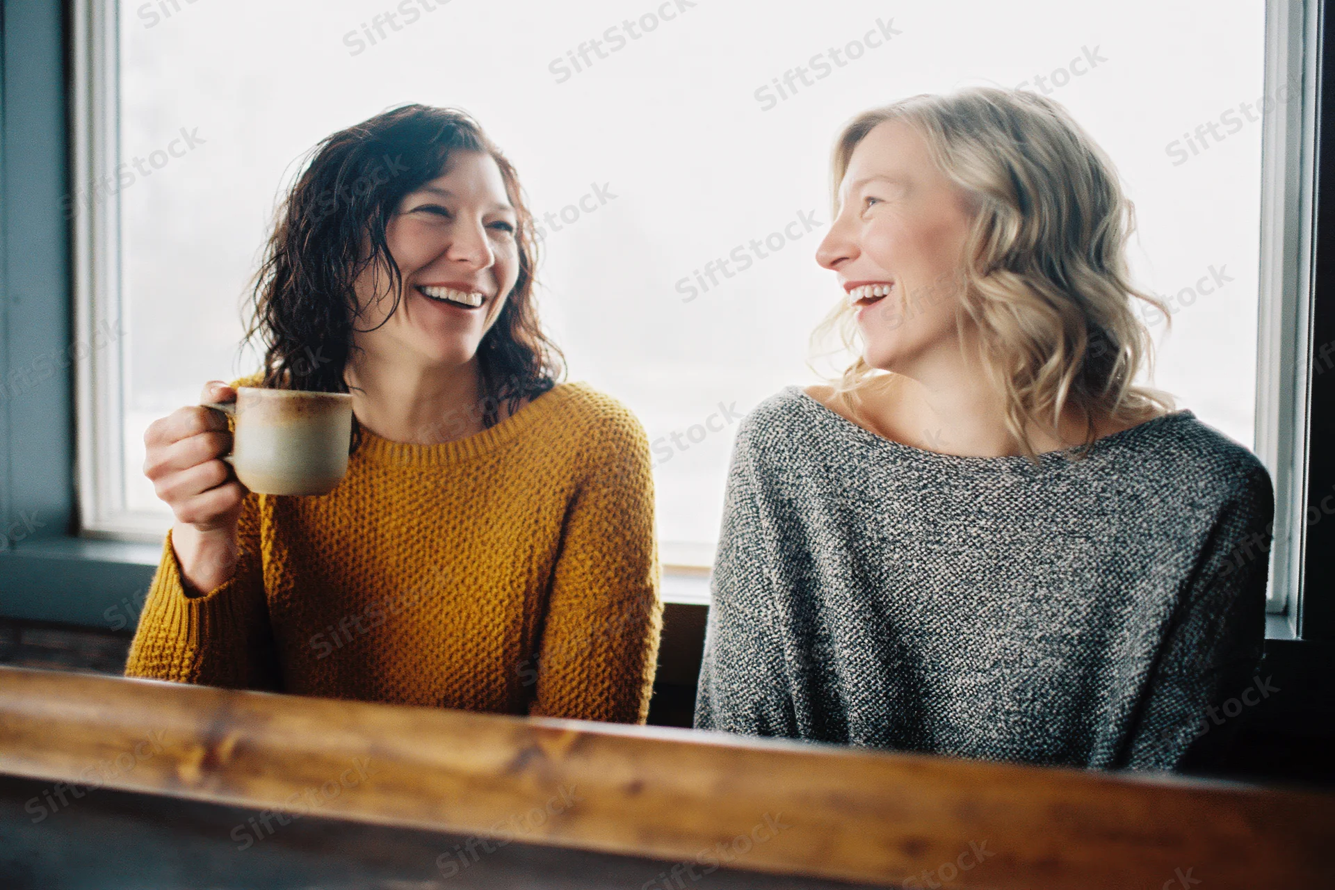 Two women laughing together at a cafe counter holding a coffee mug